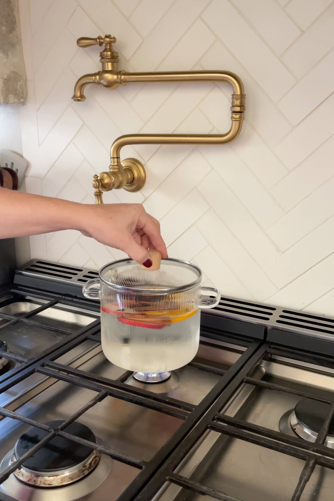 Hand placing a lid on a glass pot filled with water, apple slices, and cinnamon sticks on a stove for a Christmas Simmer Pot.